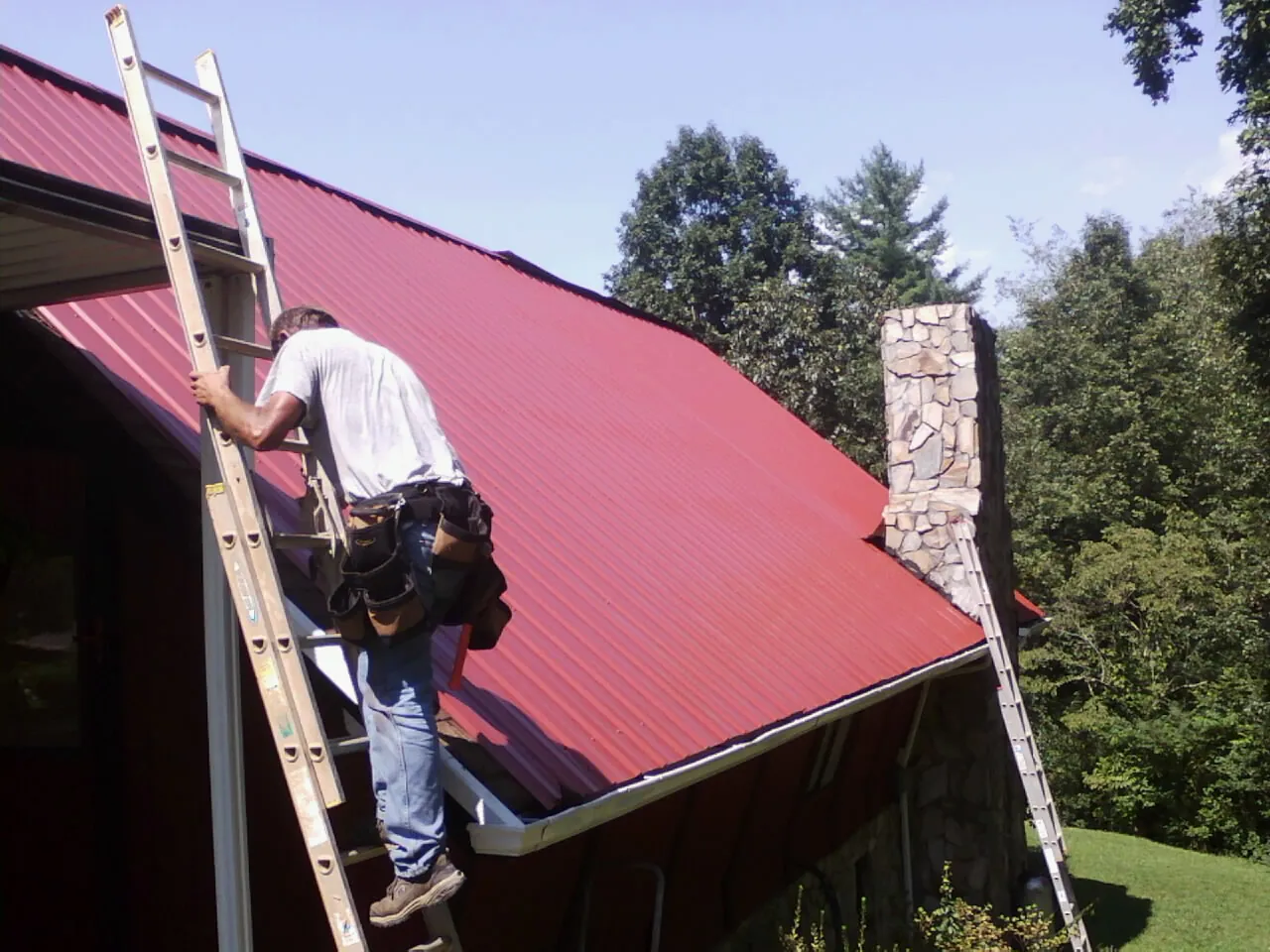 Roofer on a ladder beside a red metal roof and stone chimney — Soffit & Fascia Repair by Roofing Brothers in Bessemer